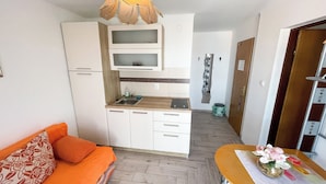 Eine kompakte Küche mit weißen Schränken, einer Mikrowelle und einem Spülbecken. Ein Holztisch mit einer Blumenvase steht daneben.