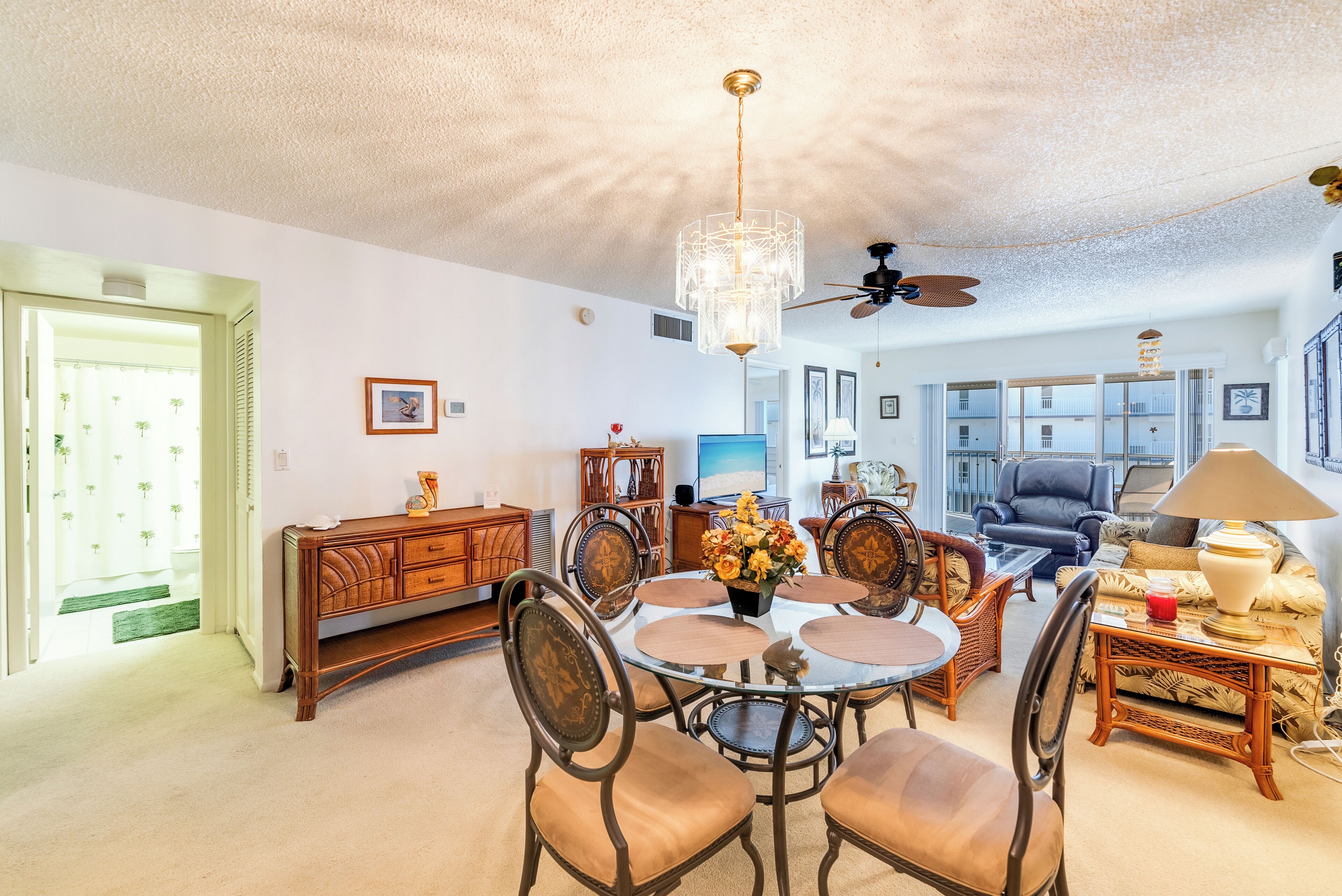 A spacious living room with a round glass table, wooden chairs, a ceiling fan, and a large window.