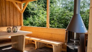 Een houten terras met een bank en tafel, een houtkachel en uitzicht op weelderige begroeiing.