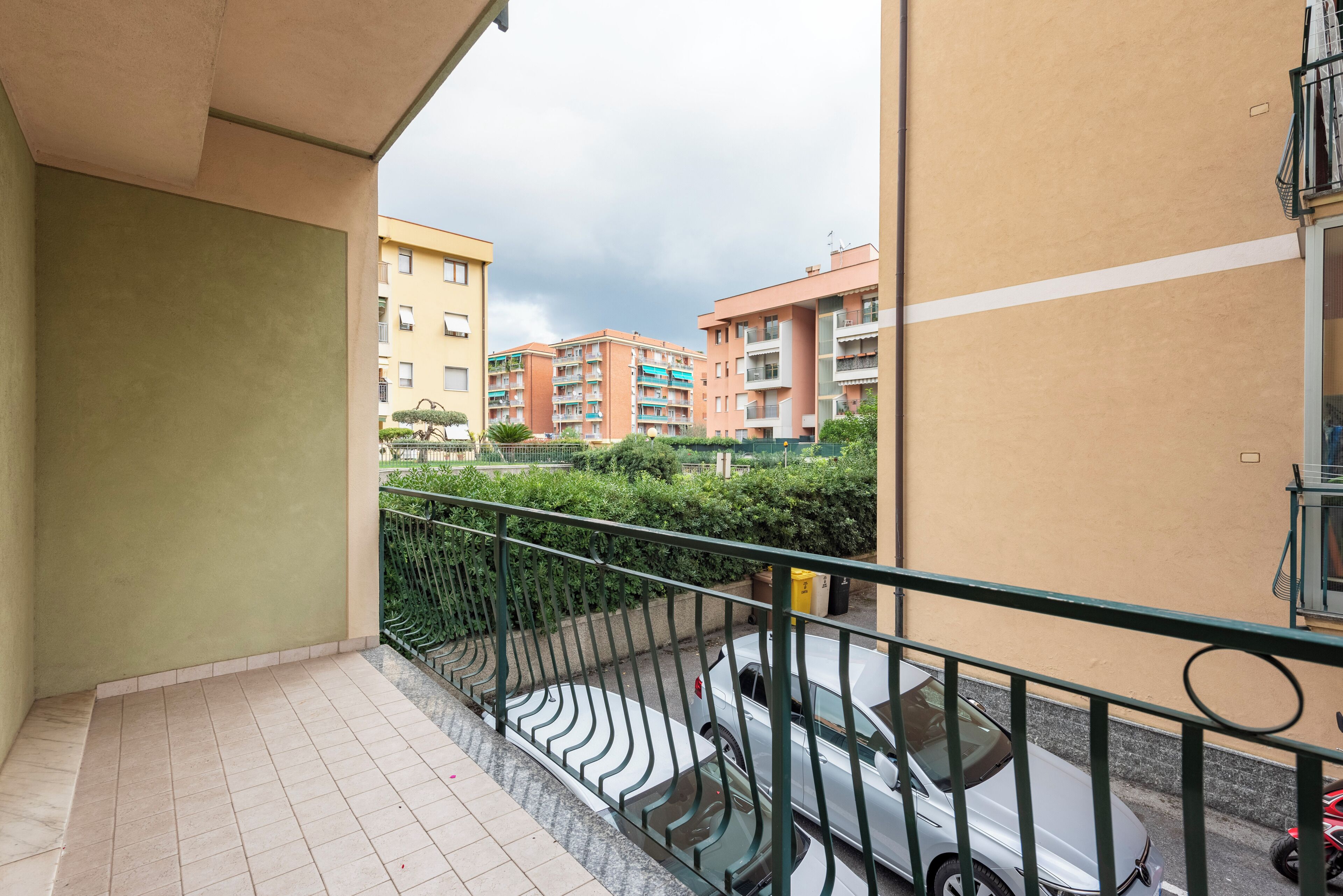 A balcony with a tiled floor, a green metal railing, and a view of multi-story buildings in the background.