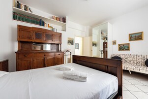 Habitación de hotel con una cama grande, un armario de madera y un sofá. Hay estanterías sobre la cama y un cuadro en la pared.