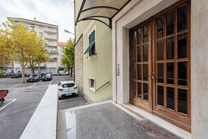 Un edificio con una puerta de madera grande, una ventana pequeña con contraventanas y un pavimento adoquinado.