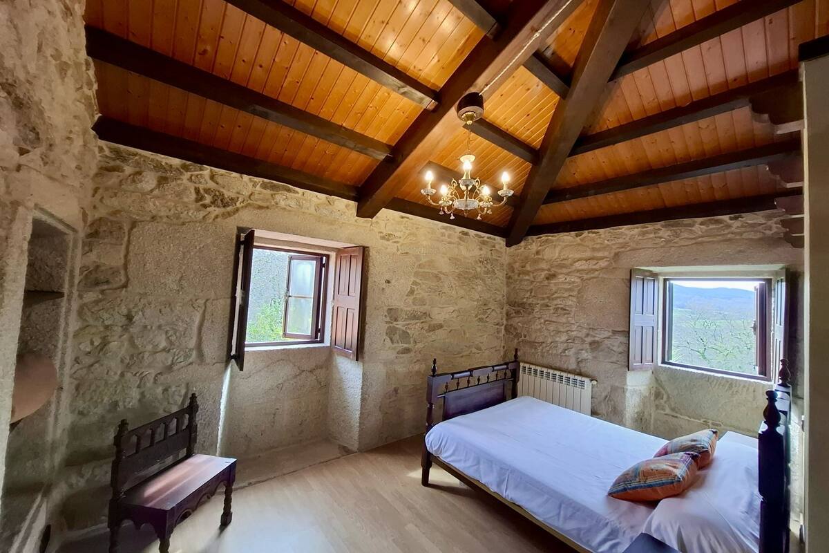 A rustic bedroom with stone walls, wooden beams, a bed with pillows, a bench, and two windows with shutters.