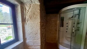 A bathroom with a stone wall, a window, and a glass-enclosed shower. - Val Fresco Suites & Relax (A Estrada)