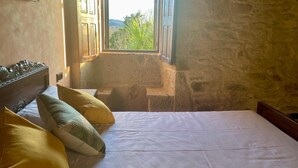 A bedroom with a bed, pillows, a wooden headboard, a small table, and a window with a view of greenery. - Val Fresco Suites & Relax (A Estrada)