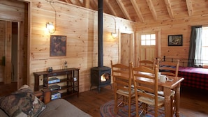 A cozy cabin interior with wooden walls, a central fireplace, a dining area, and a sofa. - ADA Cabin, Queen Bed, Double Bed, Private Bath (Greenville)