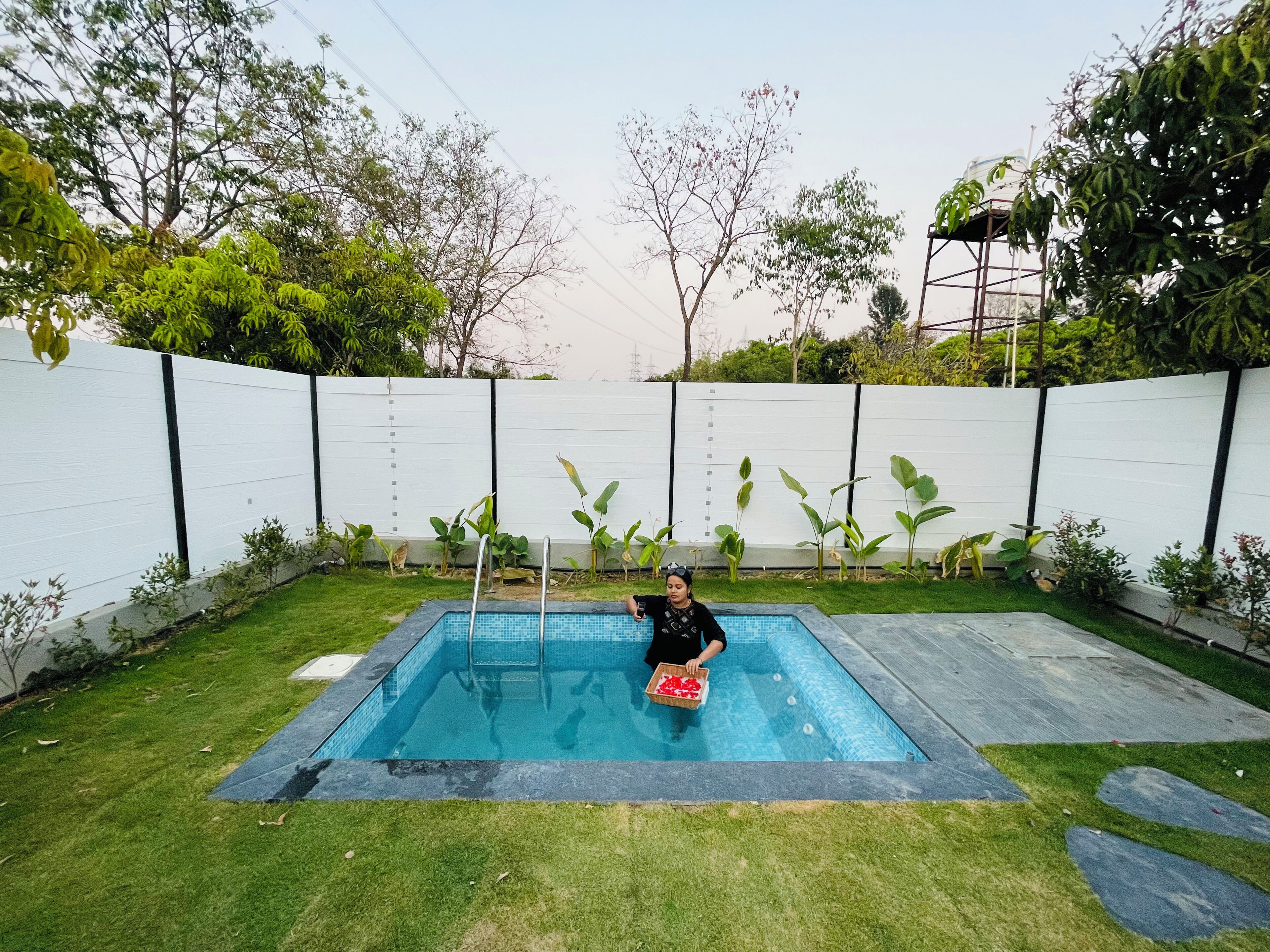 A swimming pool with a person holding a box of items, surrounded by a white fence and greenery.