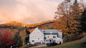 Une grande maison à deux étages, dotée d’une cheminée imposante, entourée d’arbres aux couleurs automnales et s’élevant sur un paysage vallonné.
