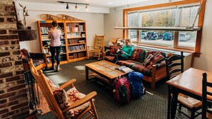 Une bibliothèque accueillante, avec des étagères remplies de livres, un coin lecture doté d’un canapé et de fauteuils, et une grande fenêtre donnant sur les voitures garées.