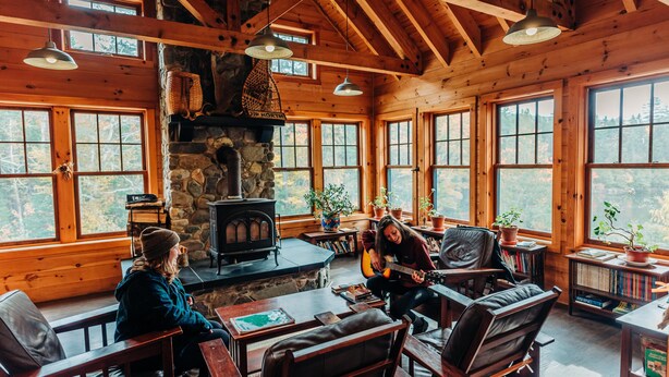A cozy cabin interior with a stone fireplace, wooden walls, and large windows offering views of the forest. - Cabin, two queen beds with shared bathroom (Greenville)