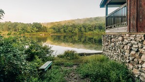 A rustic cabin with a stone foundation, a wooden deck, and a view of a calm lake surrounded by lush greenery. - Cabin, Double Bed, 2 Bunks, Shared Bathroom (Blairstown)
