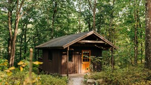 A small wooden cabin with a door and a window, surrounded by dense forest. - Cabin, Double Bed, 2 Bunks, Shared Bathroom (Blairstown)