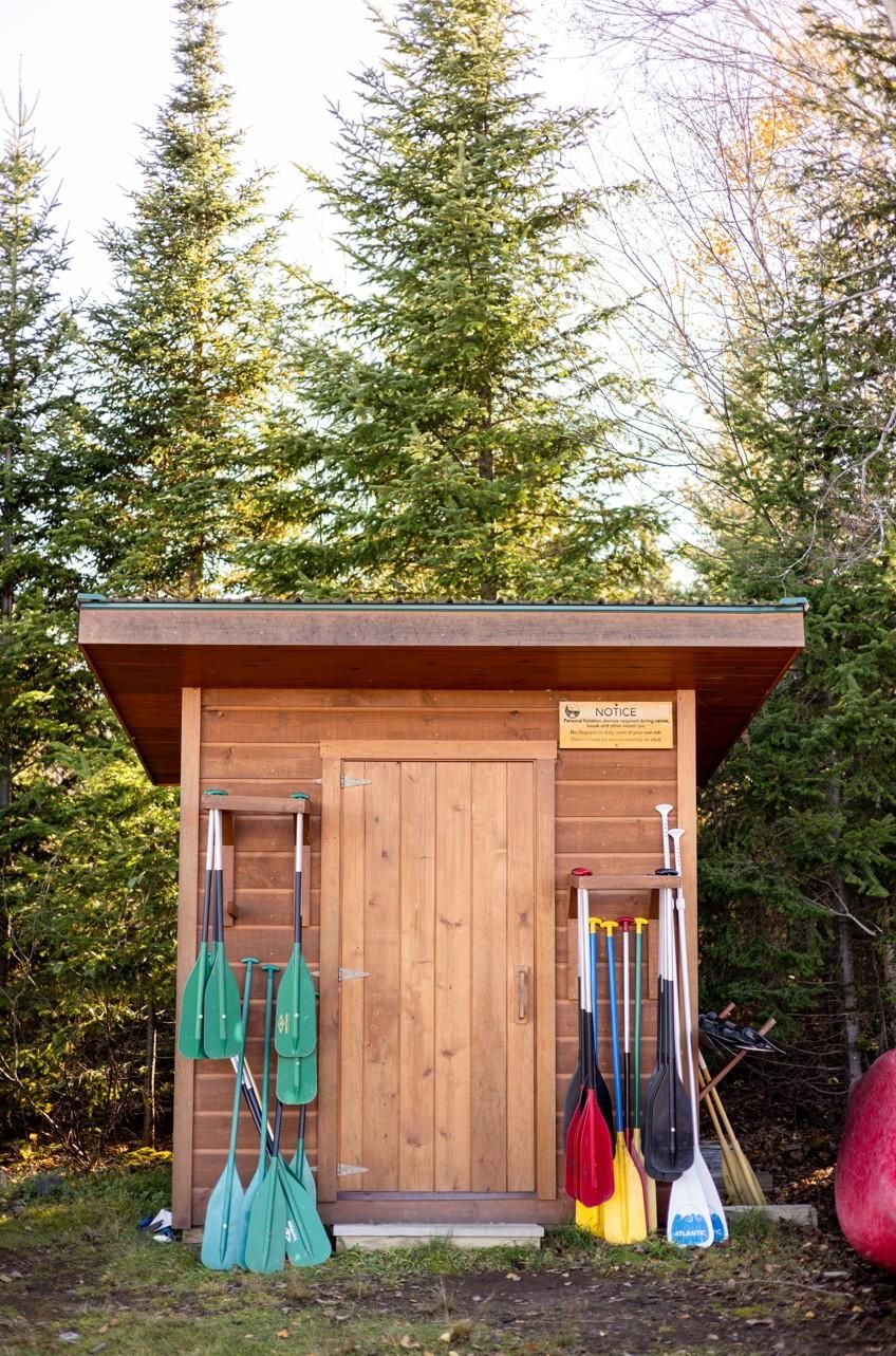 A wooden cabin with a door and a notice sign. There are several paddles and a paddleboard leaning against the cabin.