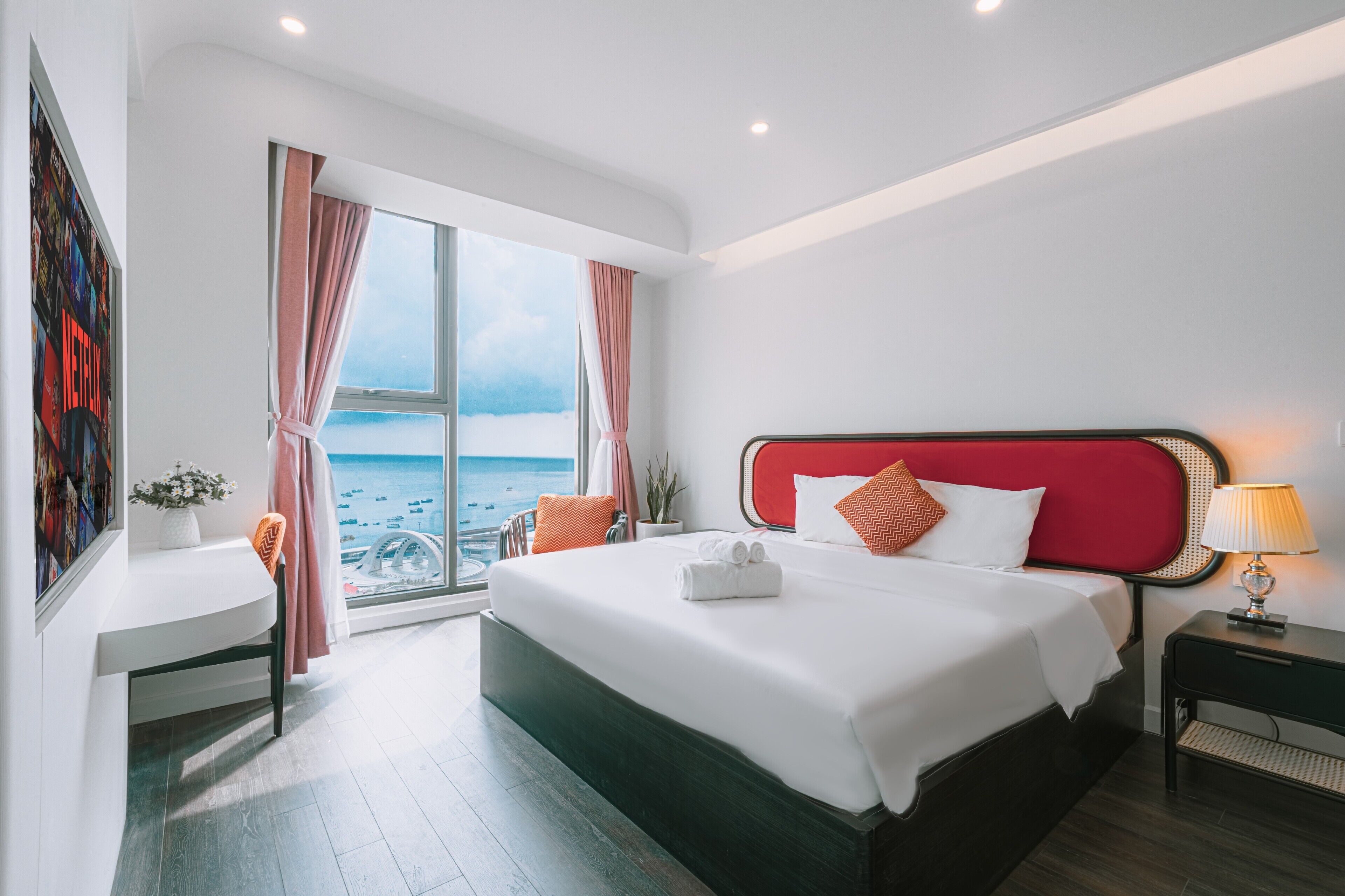 A modern hotel room with a large bed, a desk, and a view of the sea.