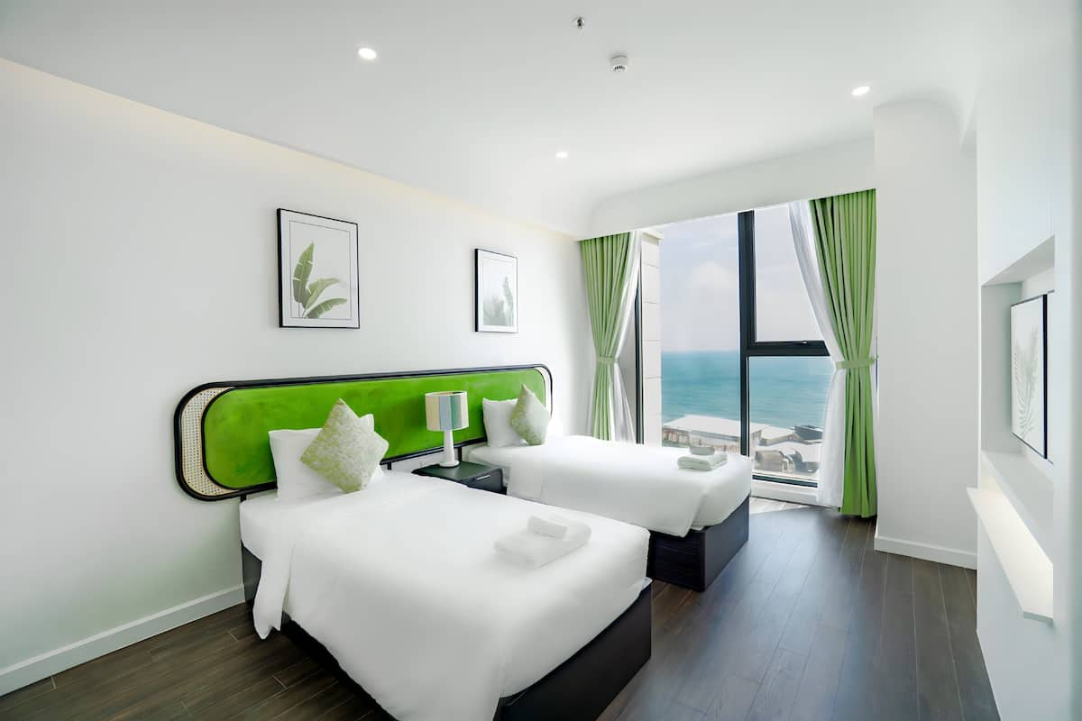 A hotel room with two beds, a large window with a view of the sea, and a green headboard.