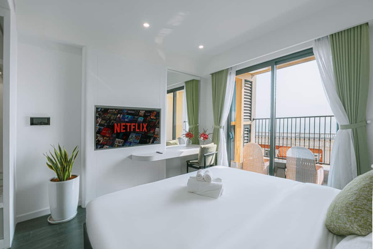 A hotel room with a bed, a desk with a computer, a potted plant, and a balcony with a view.