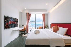 Habitación de hotel con una cama grande, un televisor mostrando un cartel de Netflix, vista al mar y un cabecero rojo.