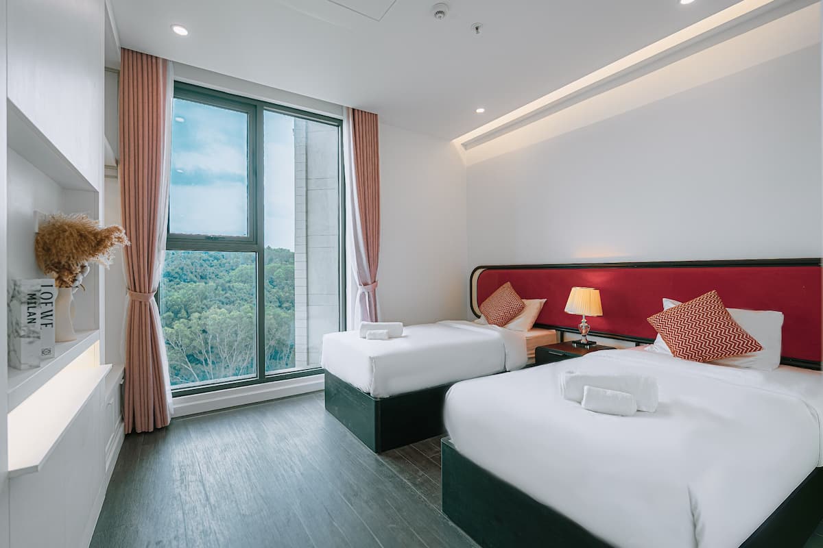 A hotel room with two beds, a large window with curtains, a red headboard, and a nightstand with a lamp.