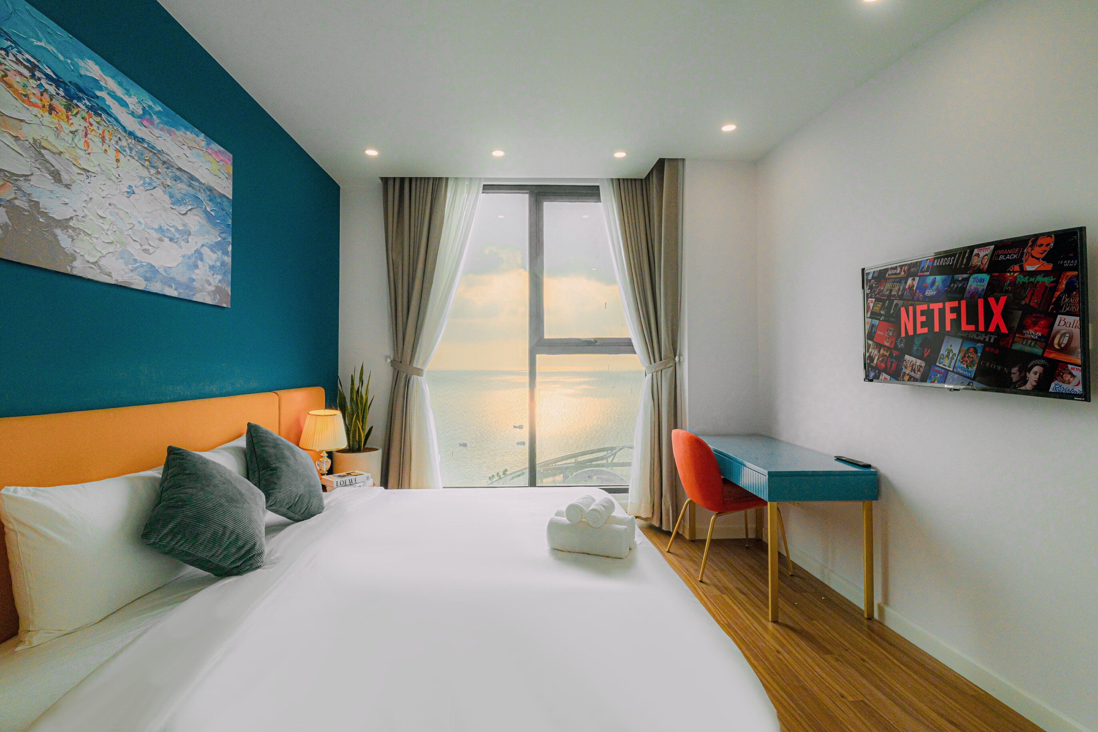 A hotel room with a bed, a desk, a chair, a large window with curtains, a wall art piece, and a wall-mounted poster.