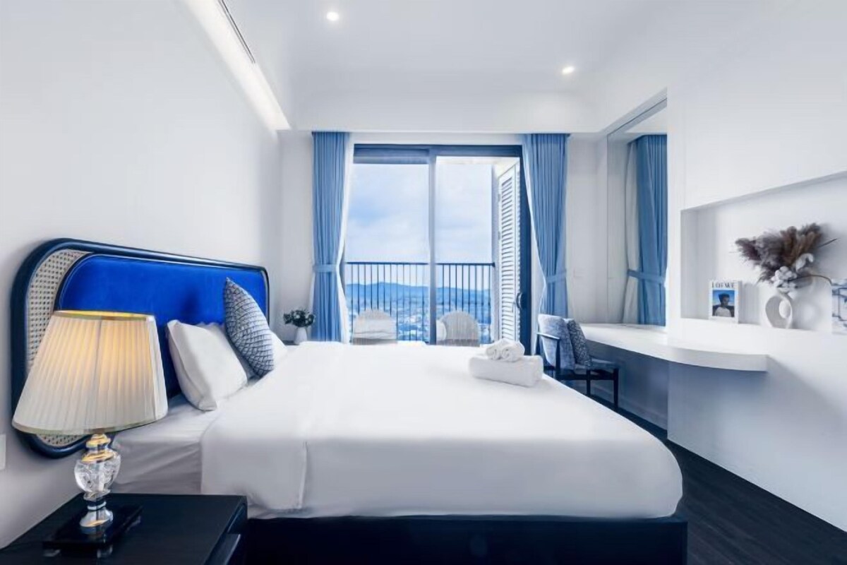 A modern hotel room with a large bed, a bedside table with a lamp, a view of the city through the window, and a small desk area.