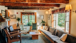A cozy living room with a fireplace, wooden furniture, and large windows offering a view of the outdoors.