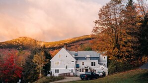 A large, two-story house with a prominent chimney, surrounded by autumn-colored trees and a hilly backdrop.