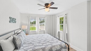 A bedroom with a bed, a ceiling fan, and large windows with curtains. - Chasing Sunsets- Close to downtown, fenced yard, pet friendly, family friendly,Sleeps 9 (Port St. Joe)