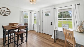 A room with a wooden dining table, chairs, a white door, and large windows with curtains. - Chasing Sunsets- Close to downtown, fenced yard, pet friendly, family friendly,Sleeps 9 (Port St. Joe)
