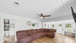 A spacious living room with a large brown leather sectional sofa, a ceiling fan, and a view of the outdoors through large windows. - Chasing Sunsets- Close to downtown, fenced yard, pet friendly, family friendly,Sleeps 9 (Port St. Joe)