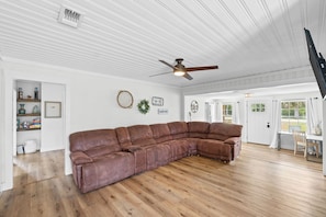 A spacious living room with a large brown leather sectional sofa, a ceiling fan, and a view of the outdoors through large windows.