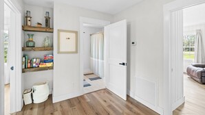A hallway with wooden flooring, white walls, and a door leading to another room. There are shelves with various items and a small rug on the floor. - Chasing Sunsets- Close to downtown, fenced yard, pet friendly, family friendly,Sleeps 9 (Port St. Joe)