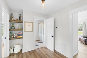 A hallway with wooden flooring, white walls, and a door leading to another room. There are shelves with various items and a small rug on the floor.