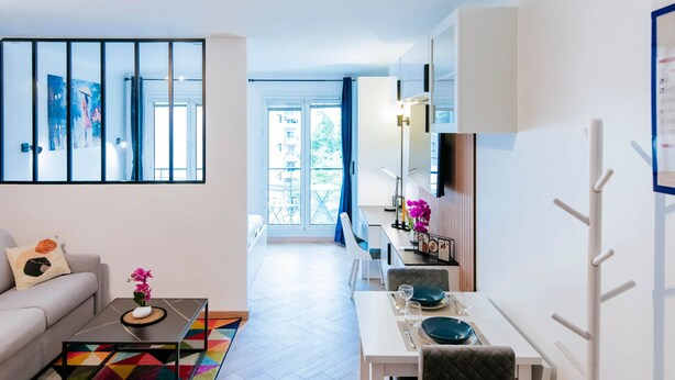 A modern living room with a sofa, coffee table, and a colorful rug. There is a dining area with a table and chairs. The room has a large window and a view of the outdoors. - Paris Porte de Versailles -Metro (Issy-les-Moulineaux)