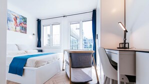 A modern bedroom with a bed, desk, chair, and a view of buildings through large windows. - Paris Porte de Versailles -Metro (Issy-les-Moulineaux)