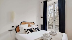 A neatly made bed with white linens and black and white pillows, a small bedside table, and a window with dark curtains.
