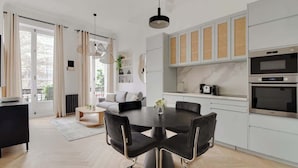 A modern kitchen with white cabinets, a black dining table with chairs, and a large window with curtains.