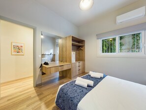 A modern bedroom with a wooden desk, a large mirror, a bed with a patterned bedspread, and a window with a view of greenery.