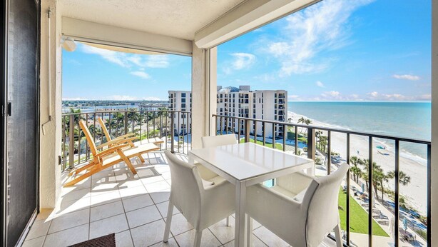 Ein Balkon mit einem weißen Tisch und Stühlen, der Blick auf einen Strand und eine Stadtansicht freigibt.