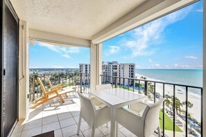 A balcony with a white table and chairs, overlooking a beach and a cityscape.