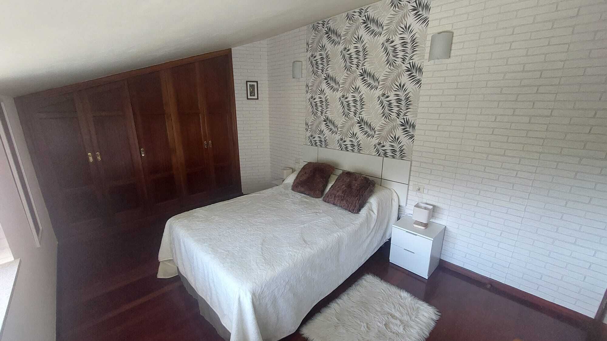 Quarto com cama, mesa de cabeceira, parede de tijolos brancos, guarda-roupa de madeira e um painel de parede estampado.