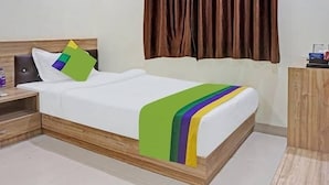 A neatly made bed with a white mattress and a colorful blanket. There is a wooden headboard and a nightstand with a bottle and a remote control. A brown curtain is partially drawn, and a wooden desk is visible in the corner. - Saalt Patliputra Hotel (Patna)