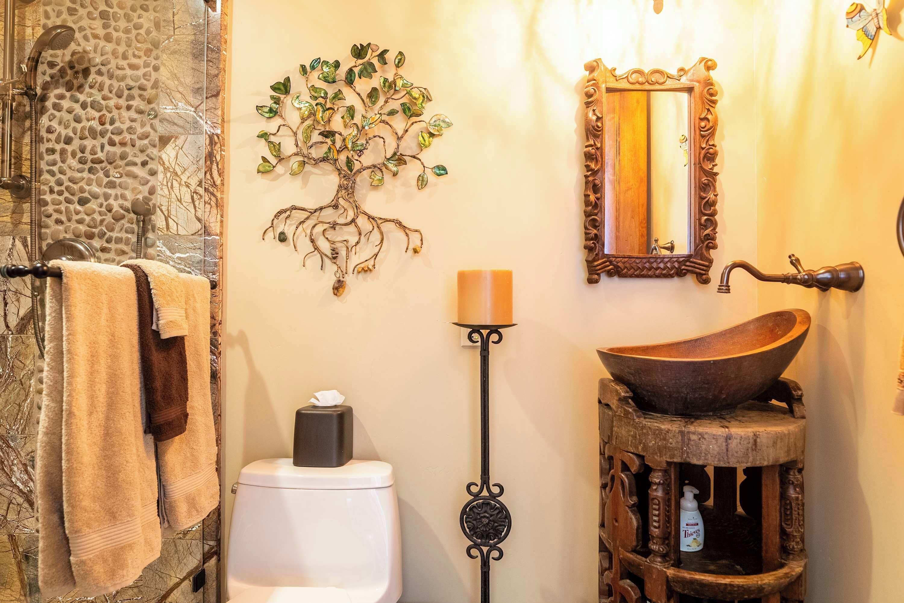 Een badkamer met een stenen douche, een decoratieve wanddecoratie van een boom, een houten wastafel en een spiegel met een sierlijke lijst.