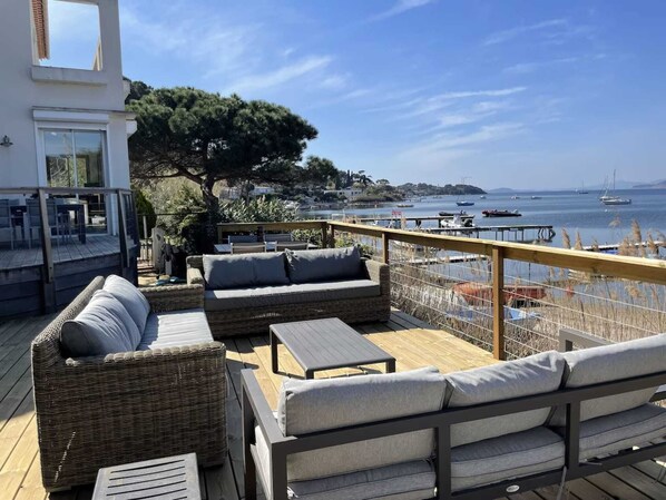 A deck with wicker furniture, a table, and a view of the sea and boats.