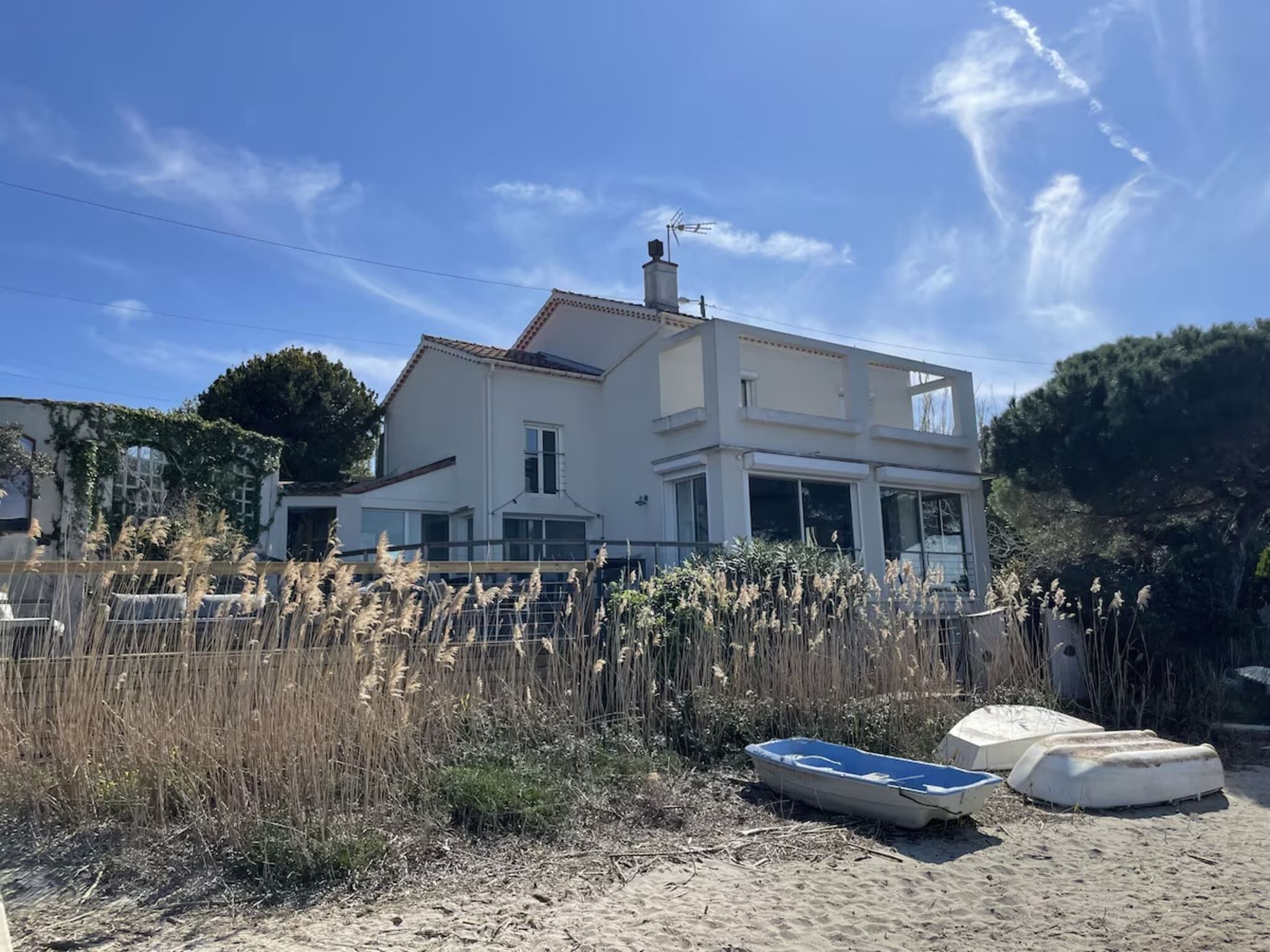 A two-story house with a large balcony, surrounded by tall grasses and a sandy area with a boat and a covered object.