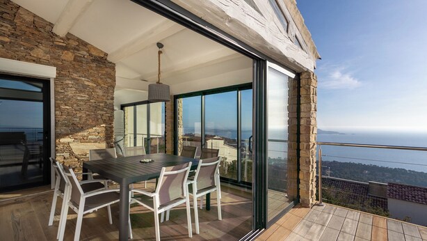 A modern outdoor dining area with a stone wall, a wooden deck, and a glass door leading to a balcony with a city view. - 180° Sea View Villa **** Truly Unique View - Hillside on the Seafront (Carqueiranne)