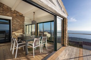 A modern outdoor dining area with a stone wall, a wooden deck, and a glass door leading to a balcony with a city view. - 180° Sea View Villa **** Truly Unique View - Hillside on the Seafront (Carqueiranne)