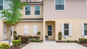 A two-story residential building with a beige exterior, white shutters, and a small front yard with various shrubs and a tree. - Magical Moments Retreat: Your Family’s Gateway to Central Florida Fun (Kissimmee)