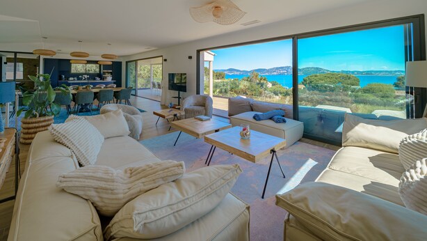 A modern living room with a large sofa, a coffee table, and a dining area with chairs and a kitchen in the background. - Villa de la Madrague Sea View - 100 m from a small beach - Giens Peninsula 12 P (Hyères)
