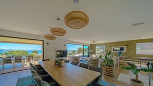 A spacious dining area with a large wooden table and modern chairs, large windows offering a view of the outdoors, and a cozy seating area with a TV and plants. - Villa de la Madrague Sea View - 100 m from a small beach - Giens Peninsula 12 P (Hyères)