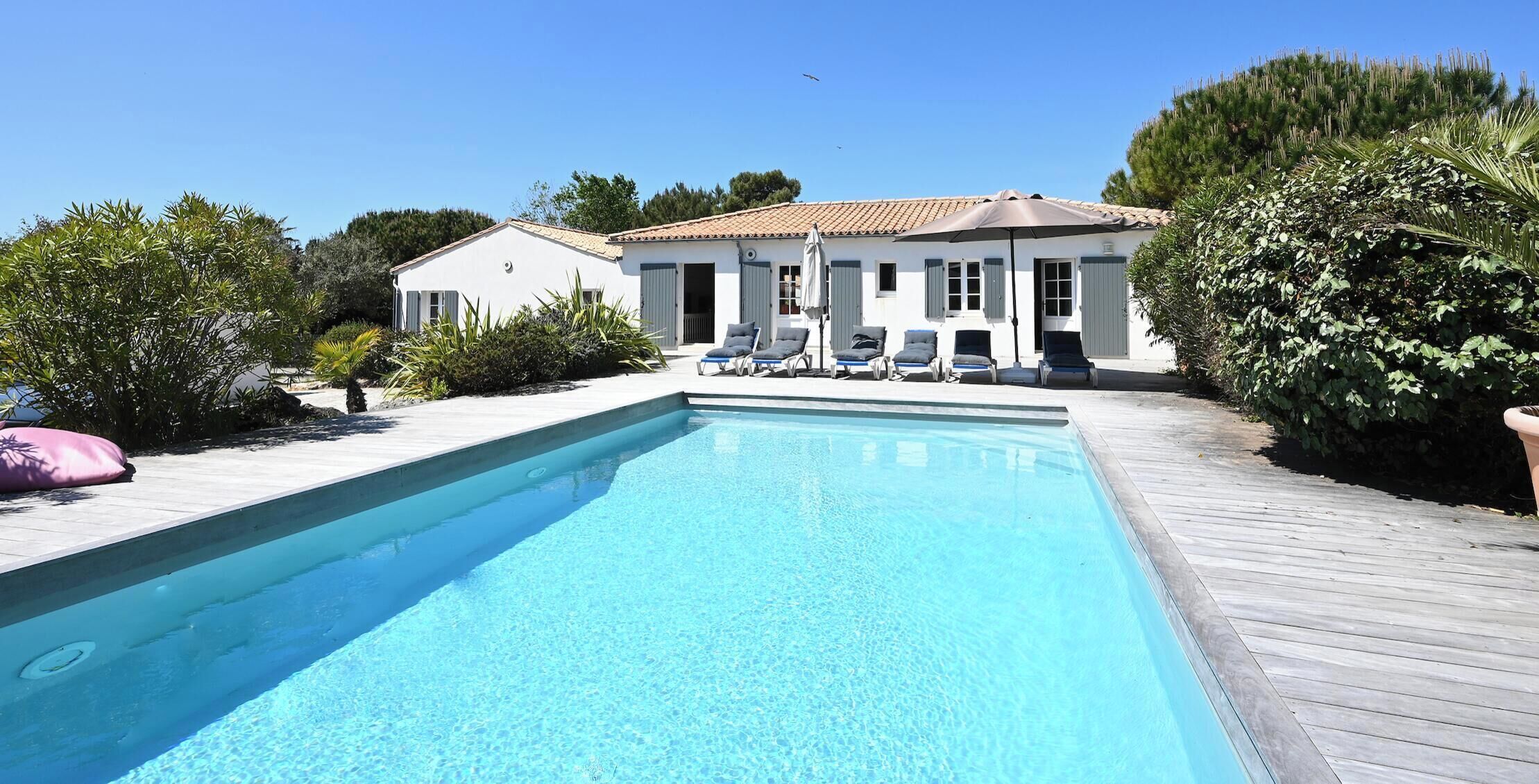 Una piscina rectangular con cubierta de madera, un edificio blanco con contraventanas y un cielo azul despejado.